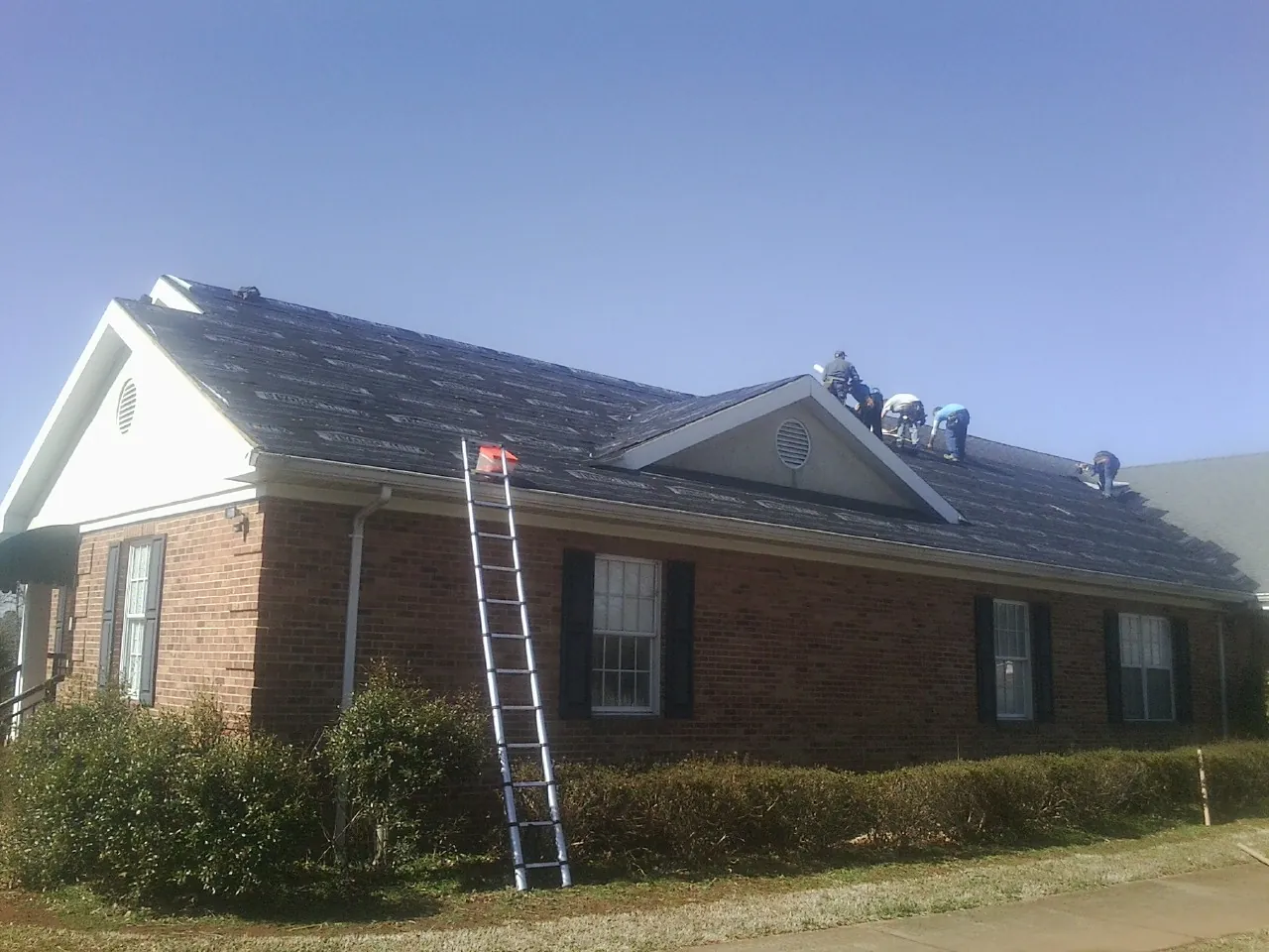 Crew tearing shingles off a brick ranch roof with a ladder set — Metal Roof Repair work by our crew in Holy Cross