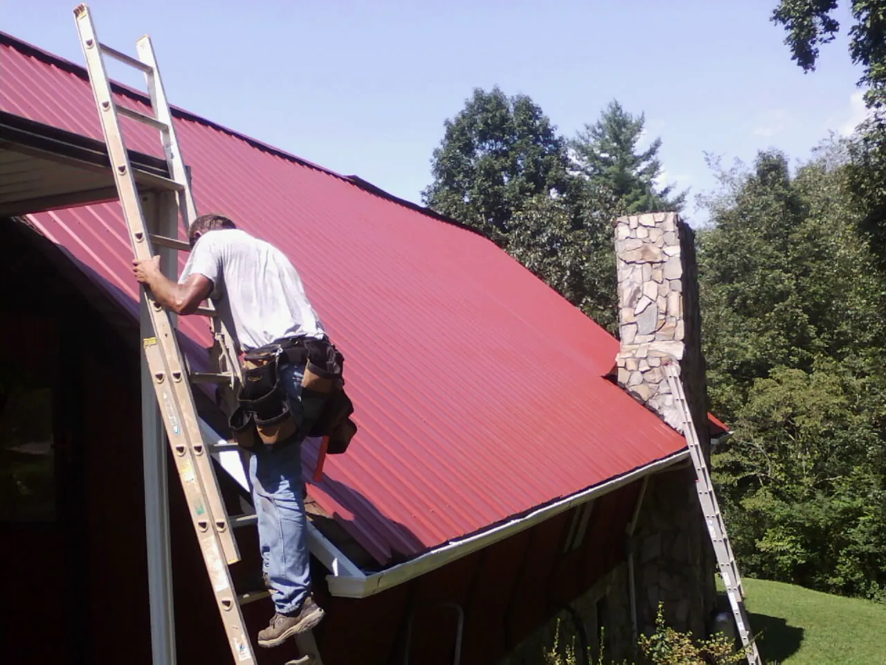 Roofer on a ladder beside a red metal roof and stone chimney — Soffit & Fascia Repair by Roofing Brothers in Holy Cross